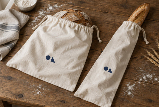 Two natural cotton bread bags with a navy logo print holding a sourdough loaf and a baguette on a bakery table